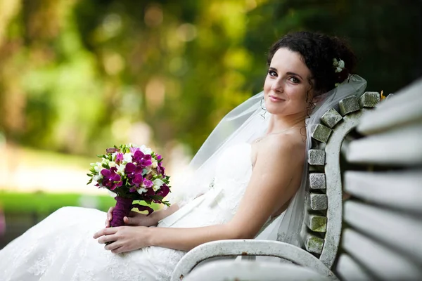 Sposa riccia tiene un mazzo di viola seduto sulla panchina — Foto Stock