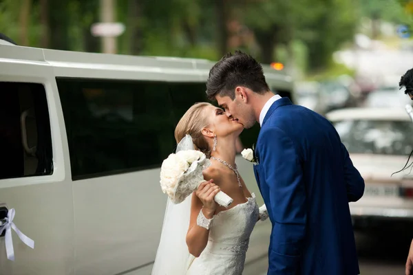 Стильная свадебная пара целуется за огромным лимузином — стоковое фото