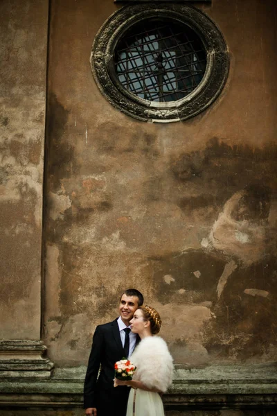Stylish wedding couple talks while standing undet the old wall w