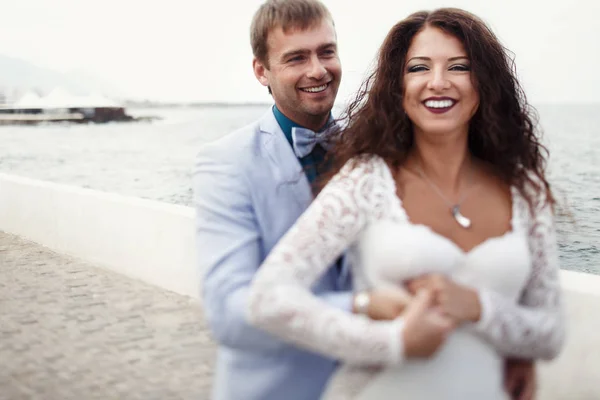 Groom segura a cintura da noiva firmemente enquanto eles posam junto ao mar — Fotografia de Stock