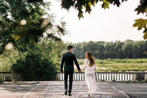 Lockigt brud har brudgummens hand medan promenader till floden — Stockfoto