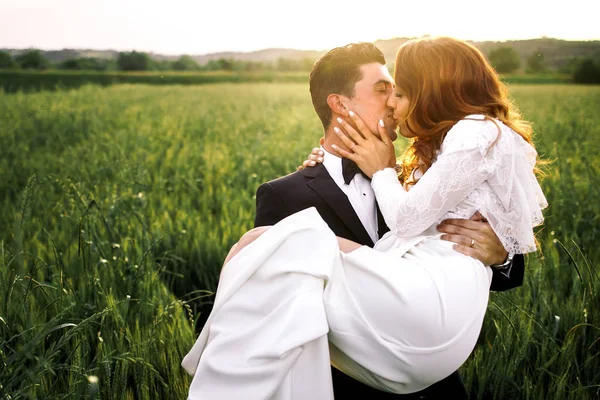 Nevěstu políbí ženicha, zatímco nosí ji prostřednictvím zelených polí — Stock fotografie