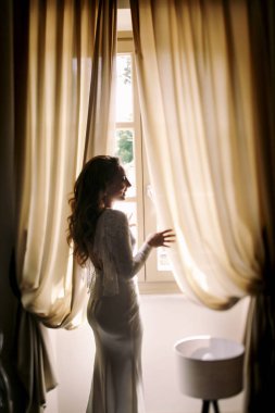 Uzun saçlı gelin bej perdeleri arasında duruyor