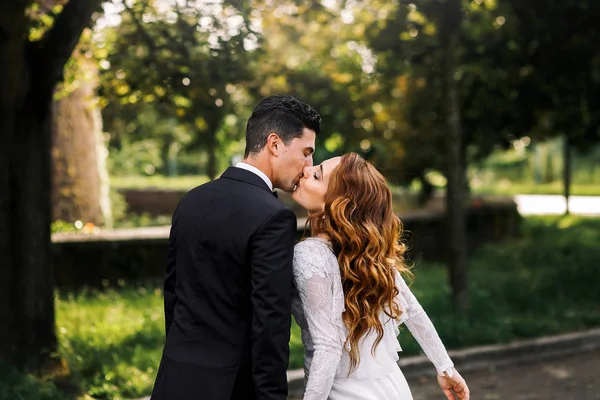 Lockige Braut lehnt sich für einen Kuss an einen Bräutigam, während sie im Par gehen — Stockfoto