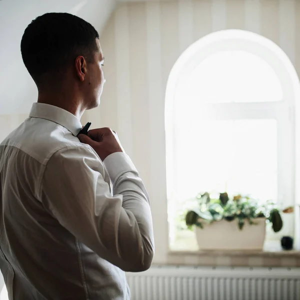 Thoughtful groom fixes bow tie standing before a window