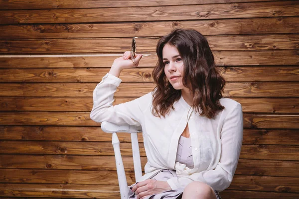 Una joven con el pelo largo, ropa blanca, en una silla y fondo de madera —  Fotos de Stock