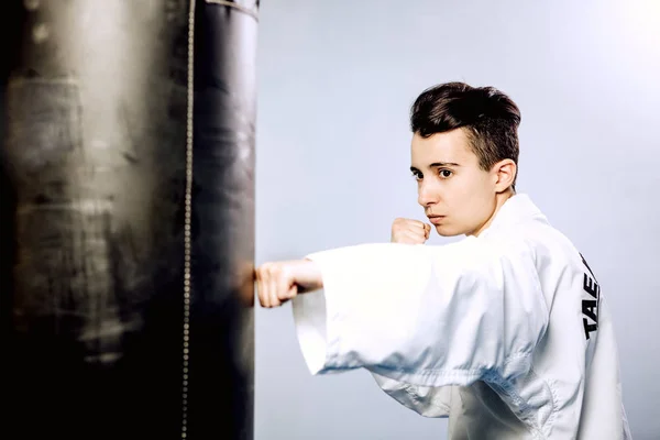 Ragazza in kimono bianco batte sacco da boxe nero su sfondo grigio. Di Tae Kwon do, karate, kick — Foto Stock