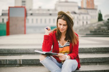 Sokak gülümseme oturan güzel, genç bir beyaz kız, sevinç, defter ile otur ve Ruhi içinde kalem. Kırmızı kazak, kot pantolon ve kahverengi deri çanta