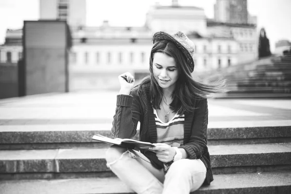 Una hermosa y joven chica caucásica sentada en la calle sonríe, la alegría, siéntate con el cuaderno y la pluma en Ruhi. En el suéter rojo, jeans y bolso de cuero marrón —  Fotos de Stock