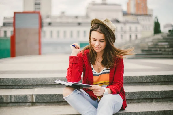 Sokak gülümseme oturan güzel, genç bir beyaz kız, sevinç, defter ile otur ve Ruhi içinde kalem. Kırmızı kazak, kot pantolon ve kahverengi deri çanta