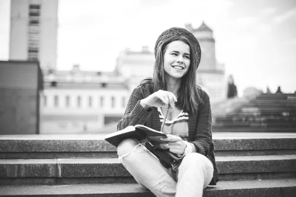 Uma linda menina caucasiana sentada na rua sorri, a alegria, senta-se com caderno e caneta em Ruhi. Na camisola vermelha, jeans e bolsa de couro marrom — Fotografia de Stock