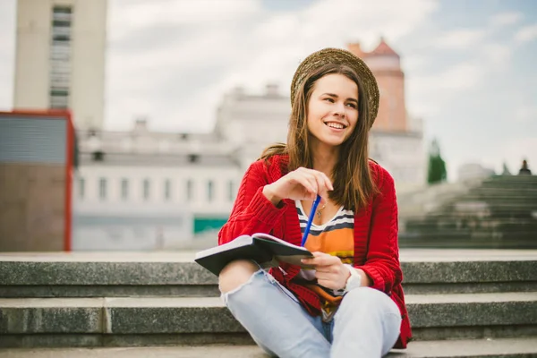 Uma linda menina caucasiana sentada na rua sorri, a alegria, senta-se com caderno e caneta em Ruhi. Na camisola vermelha, jeans e bolsa de couro marrom — Fotografia de Stock