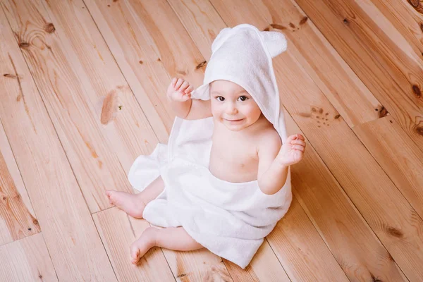 Bébé garçon aux yeux marron est âgé de cinq mois enveloppé dans une serviette blanche avec des oreilles sur fond en bois  . — Photo