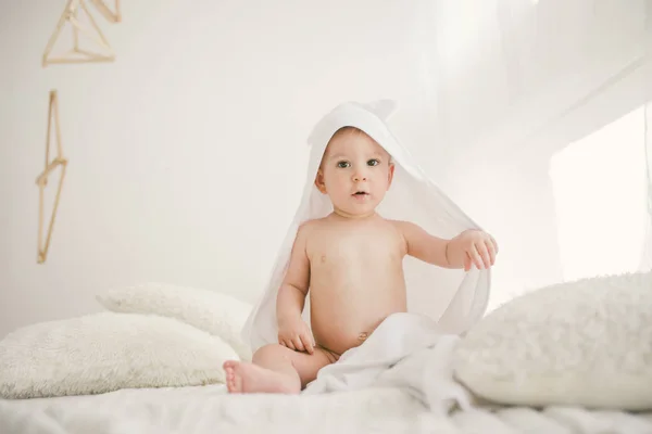 Красивый улыбающийся новорожденный мальчик, покрытый белым бамбуковым полотенцем с веселыми ушами. Сидя на белом трикотаже, шерстяной клетчатый яркий интерьер. Выход из окна — стоковое фото