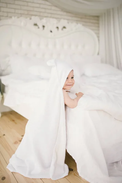 Bebé recién nacido bebé de cinco meses en el dormitorio junto a una gran cama blanca en el suelo de madera envuelto en una toalla de bambú blanco . — Foto de Stock