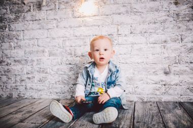 Şık bir çocuk, bir çocuk, bir yaşında, bir ahşap zemin üzerinde ve bir tuğla duvar zemin üzerine otur. Junky bir ceket, junks ve beyaz T-shirt giymiş. Sarışın