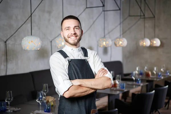 A young, smiling waiter in a restaurant, standing next to the tables with a glass of wine. He wears an apron, looks confidently, folded arms over his chest — Stock Photo, Image