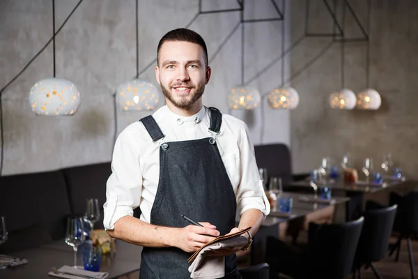 Un joven camarero sonriente en un restaurante, de pie junto a las mesas con una copa de vino. Vestido con un delantal, tomará una orden sosteniendo un cuaderno y una pluma — Foto de Stock