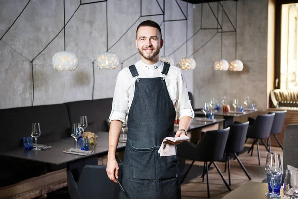 Un joven camarero sonriente en un restaurante, de pie junto a las mesas con una copa de vino. Vestido con un delantal, tomará una orden sosteniendo un cuaderno y una pluma —  Fotos de Stock