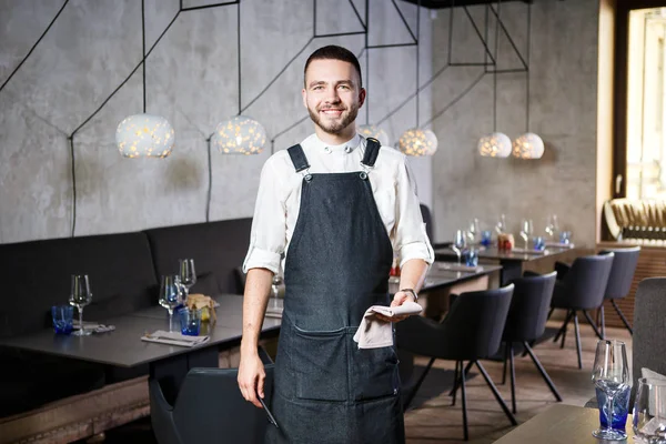 Een jonge, lachende ober in een restaurant, permanent naast de tabellen met een glas wijn. Gekleed in een schort, vindt een order houden van een notebook en pen — Stockfoto