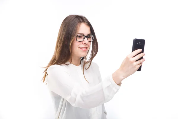 Bella ragazza in occhiali e una camicia bianca scatta una foto di se stessa sul telefono della fotocamera, selfie su sfondo bianco isolato — Foto Stock