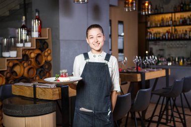 Ganson, subaylar, güzel deushka bir gülümseme ve çalışma formu önlük toplanan saç Restoran ayakta servered ahşap masa yakınındaki bir sipariş kabul eder.