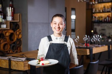 Ganson, subaylar, güzel deushka bir gülümseme ve çalışma formu önlük toplanan saç Restoran ayakta servered ahşap masa yakınındaki bir sipariş kabul eder.