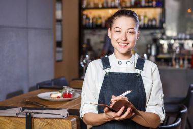 Ganson, subaylar, güzel deushka bir gülümseme ve çalışma formu önlük toplanan saç Restoran ayakta servered ahşap masa yakınındaki bir sipariş kabul eder.