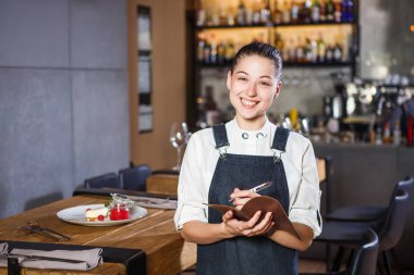 Ganson, subaylar, güzel deushka bir gülümseme ve çalışma formu önlük toplanan saç Restoran ayakta servered ahşap masa yakınındaki bir sipariş kabul eder.