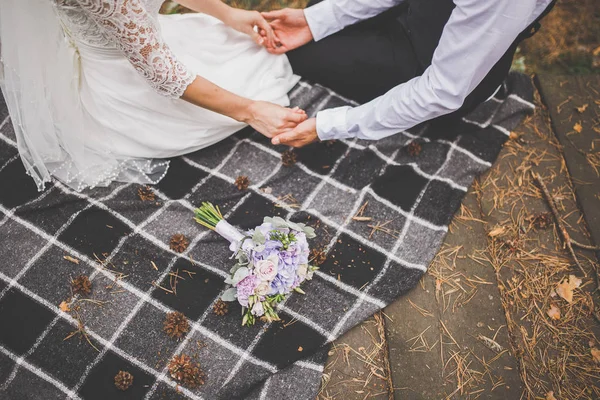 La novia y el novio están sentados en una tela a cuadros tomados de la mano. Un ramo de bodas cerca — Foto de Stock