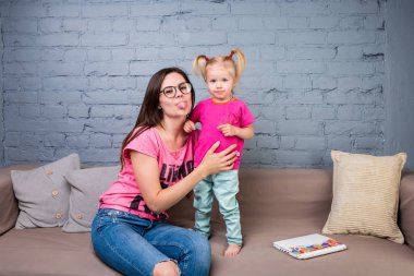 Anne ve bebek oynamak ve eğlenmek kanepede odasında. Onlar parlak giysilerle giyindin. Çekici, şık ve genç ailesi