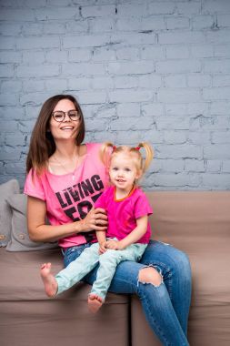 Anne ve bebek oynamak ve eğlenmek kanepede odasında. Onlar parlak giysilerle giyindin. Çekici, şık ve genç ailesi