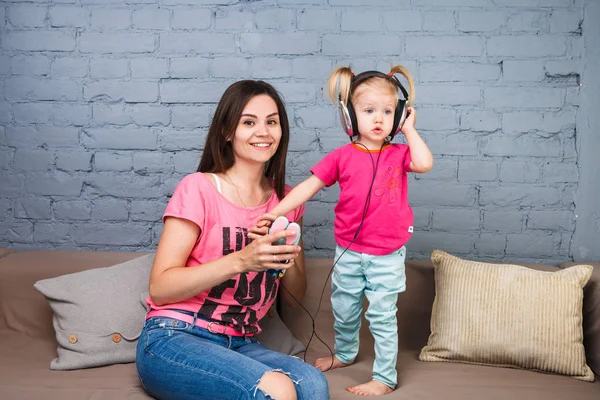 Maman et fille écoutent de la musique dans de gros écouteurs mis sur leur tête, assis sur le canapé. Tient le téléphone . — Photo