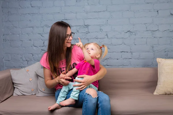 Anne ve bebek oynamak ve eğlenmek kanepede odasında. Onlar parlak giysilerle giyindin. Çekici, şık ve genç ailesi