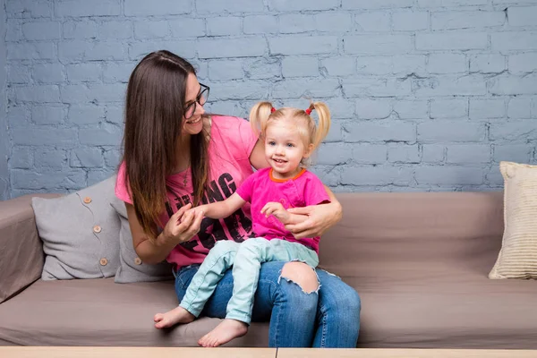Anne ve bebek oynamak ve eğlenmek kanepede odasında. Onlar parlak giysilerle giyindin. Çekici, şık ve genç ailesi — Stok fotoğraf