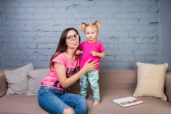 Anne ve bebek oynamak ve eğlenmek kanepede odasında. Onlar parlak giysilerle giyindin. Çekici, şık ve genç ailesi