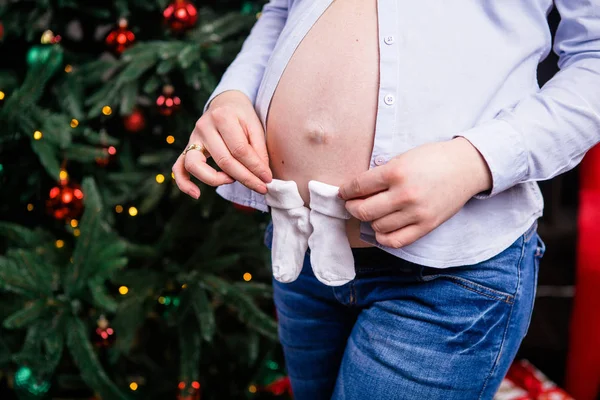 Μια έγκυος γυναίκα που κρατά στα χέρια των παιδιών της κάλτσες, καλτσάκια στο φόντο ενός χριστουγεννιάτικου δέντρου. Ντυμένος με ένα πουκάμισο και παλιατζούρες. κινηματογράφηση σε πρώτο πλάνο — Φωτογραφία Αρχείου