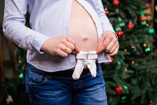 A pregnant woman holds in her hands children's socks, booties on the background of a Christmas tree. Dressed in a shirt and junks. closeup — Stock Photo, Image