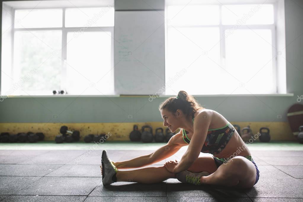 Sporty young girl doing warm-up, stretching muscles on the floor in gymnasium in backlight from window