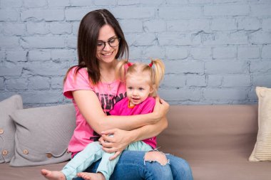 Anne ve bebek oynamak ve eğlenmek kanepede odasında. Onlar parlak giysilerle giyindin. Çekici, şık ve genç ailesi