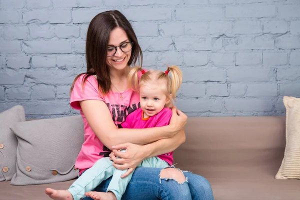 Anne ve bebek oynamak ve eğlenmek kanepede odasında. Onlar parlak giysilerle giyindin. Çekici, şık ve genç ailesi