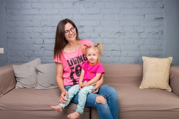 Anne ve bebek oynamak ve eğlenmek kanepede odasında. Onlar parlak giysilerle giyindin. Çekici, şık ve genç ailesi — Stok fotoğraf