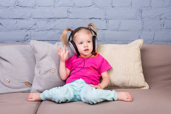 Kanepe yastık ile oturan ve başının üstüne koy büyük kulaklıklar müzik bebek kız. — Stok fotoğraf