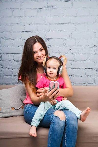 ママと娘ソファーに座って自分の頭に置く大きなヘッドホンで音楽を聴きます。携帯電話を保持しています。. — ストック写真