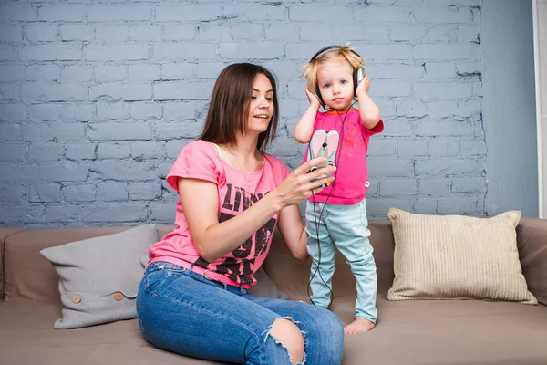 Mom and daughter listen to music in big headphones put on their head, sitting on the sofa. Holds the phone.