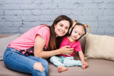 Anne ve kızı iki yıl kafası üzerinde kulaklık ile müzik dinlemek. Bir mavi tuğla duvara odasında Kanepeye otur. Kucaklama ve gülümseme