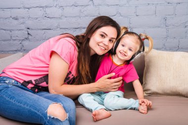 Anne ve kızı iki yıl kafası üzerinde kulaklık ile müzik dinlemek. Bir mavi tuğla duvara odasında Kanepeye otur. Kucaklama ve gülümseme
