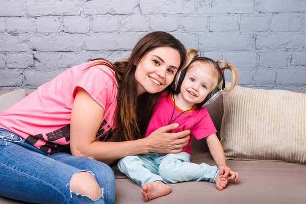 Anne ve kızı iki yıl kafası üzerinde kulaklık ile müzik dinlemek. Bir mavi tuğla duvara odasında Kanepeye otur. Kucaklama ve gülümseme — Stok fotoğraf