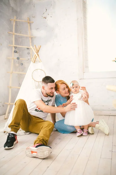 Een gelukkig gezin van drie. Vader, moeder, kind één jaar oud meisje in jurk spelen, lachen, glimlachen in lichte kamer. Zonnige — Stockfoto