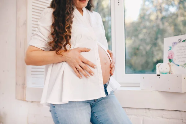 W ciąży młoda matka trzyma ręce na jej brzuch. Szczelnie-do góry. Ubrany w białą koszulę i niebieskie dżinsy — Zdjęcie stockowe